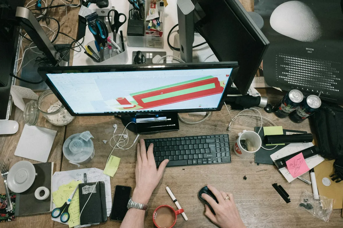 A working desk seen from above, hands at a keyboard surrounded by notes, cables and stationery