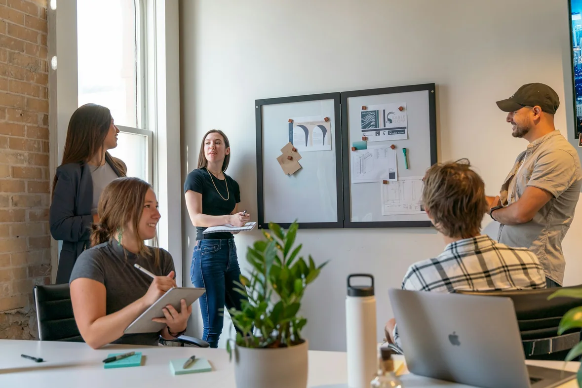 Colleagues gathered around a pinboard of printed pages in a bright office, discussing the work in progress