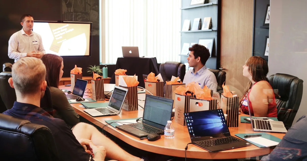 A presenter standing beside a screen addressing colleagues seated around a boardroom table with open laptops
