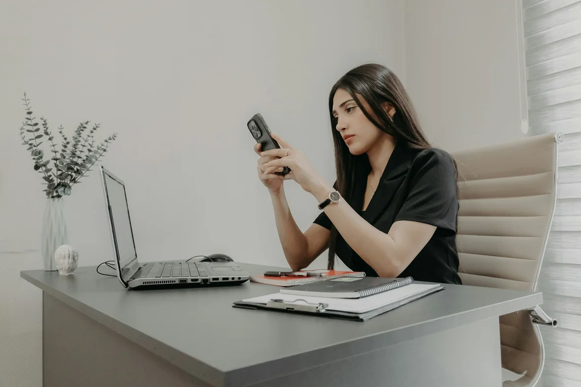 A woman using her phone at a desk beside a laptop