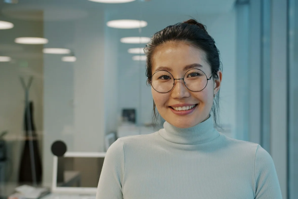 A professional portrait of a woman wearing glasses, an office visible softly behind her