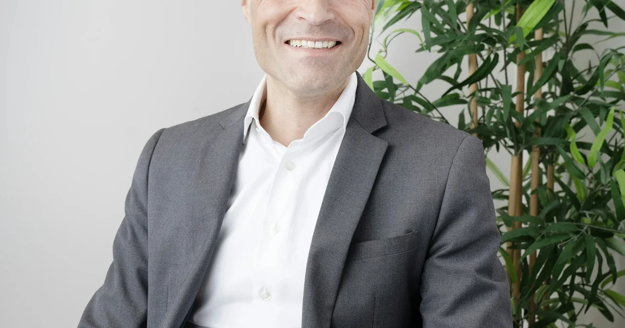 A professional portrait of a man in a grey suit seated indoors, smiling towards the camera