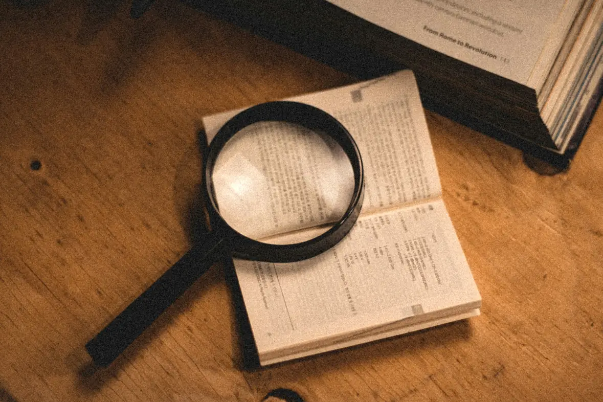A magnifying glass resting on an open book beside a stack of older volumes on a wooden desk