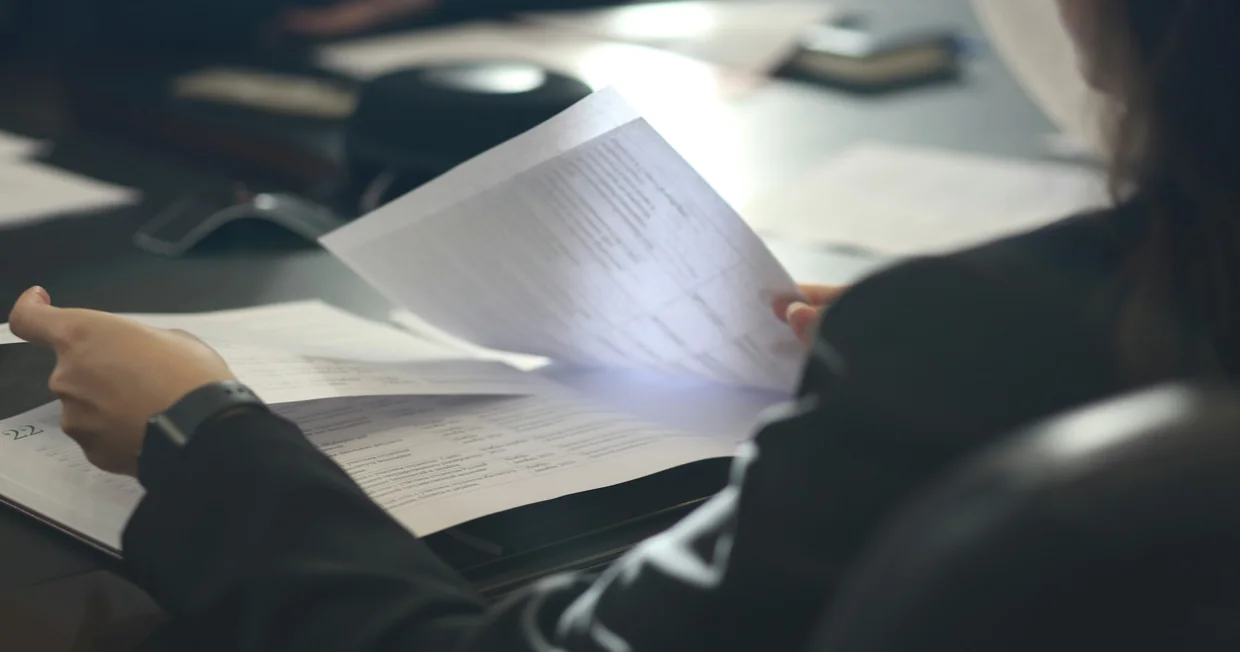 Hands turning the pages of a printed document at a meeting table, papers spread out under low light