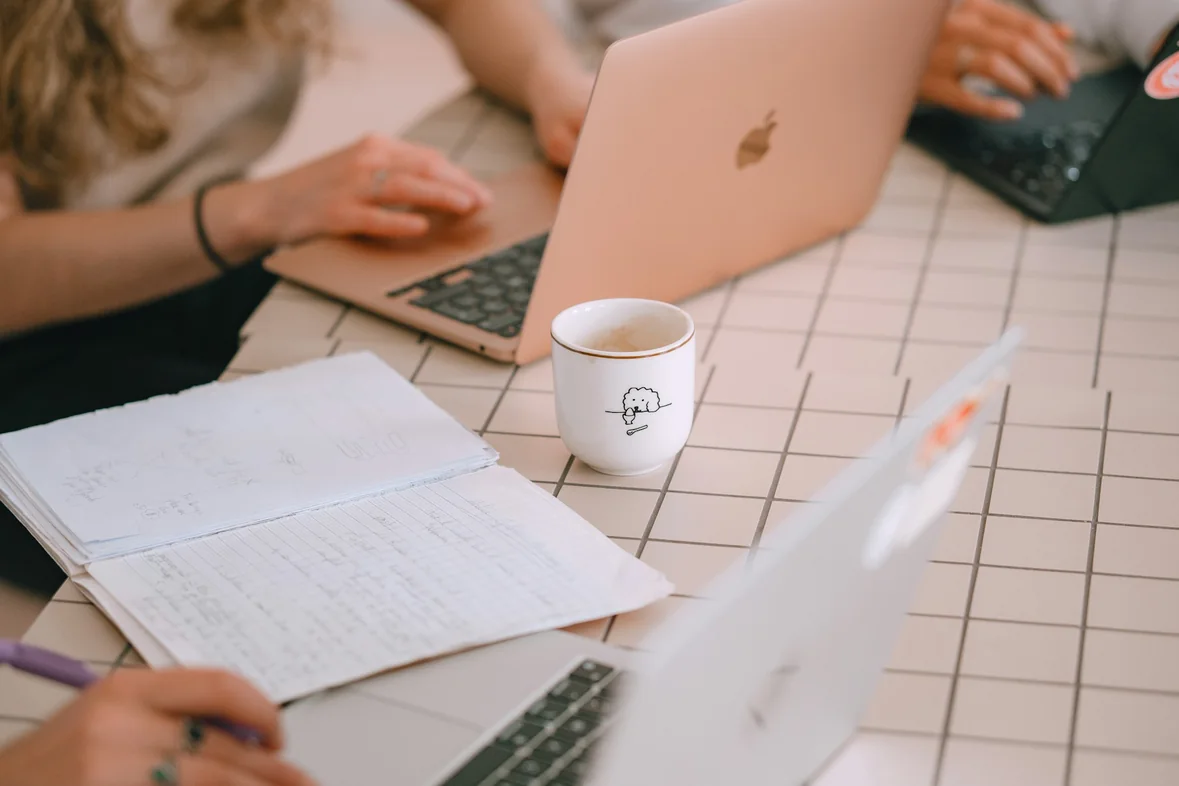 Three people working together at a table with open laptops, a notebook of handwritten notes and a coffee cup between them