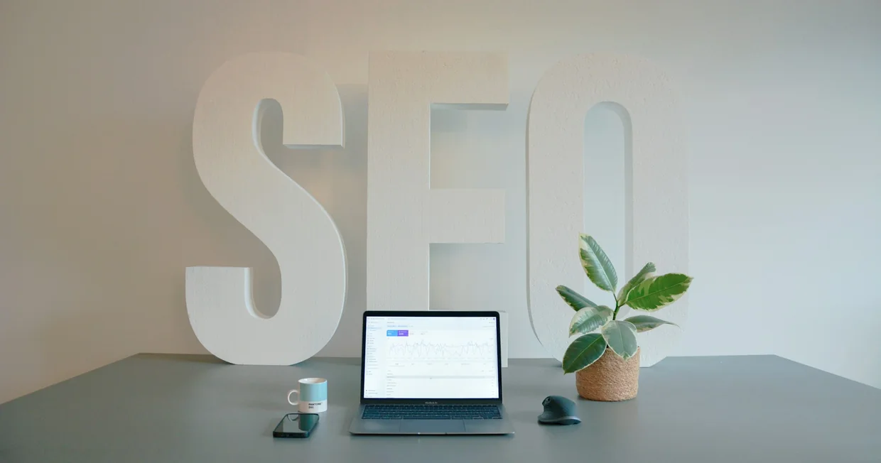 Large white three-dimensional letters spelling SEO standing against a pale wall, with an open laptop showing a traffic dashboard on the desk in front of them