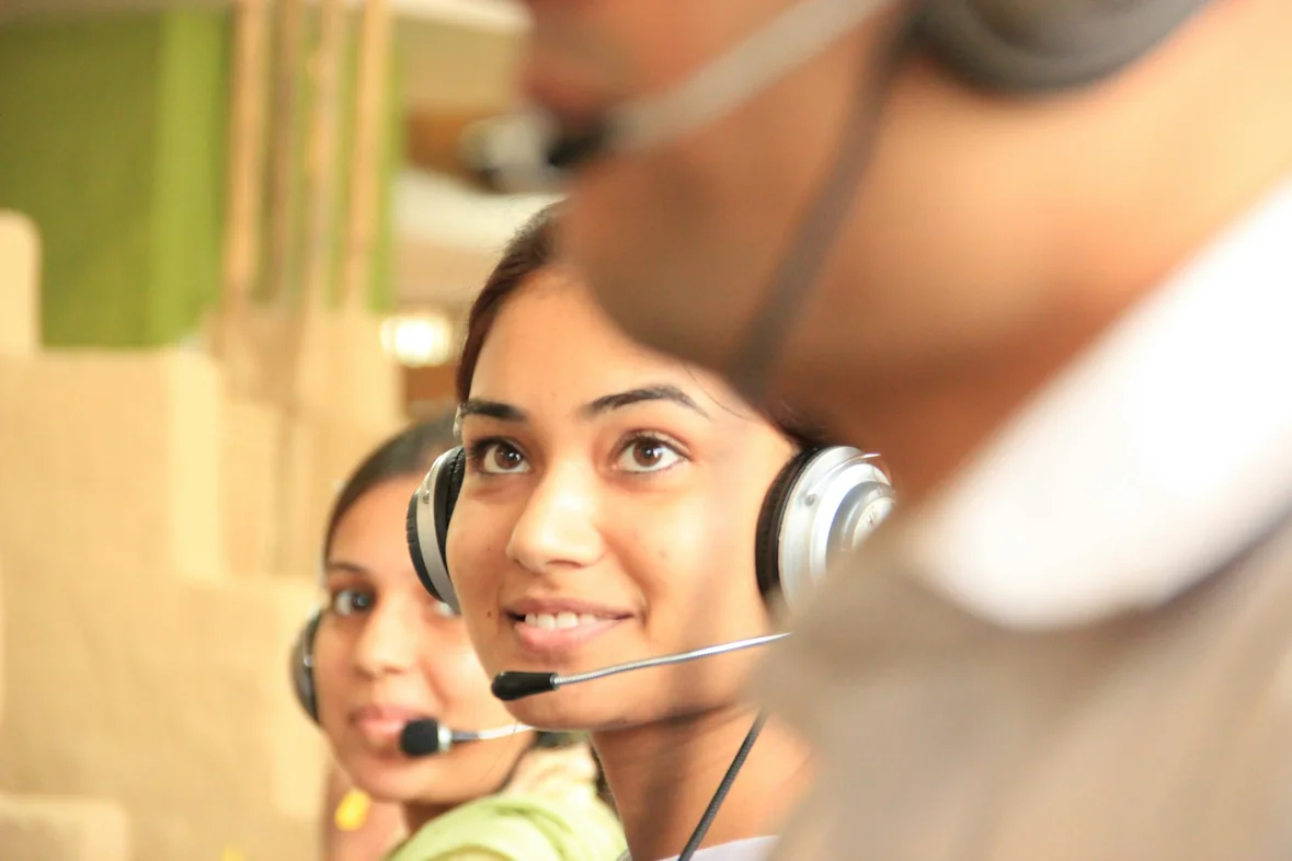 Two colleagues in headsets at a shared workstation, one smiling mid-conversation