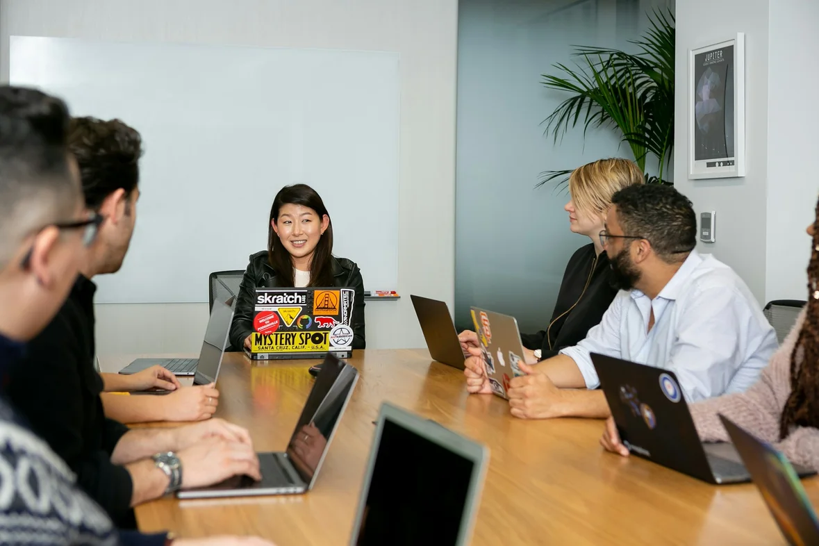 A meeting in a conference room, several people with open laptops facing a colleague leading the discussion