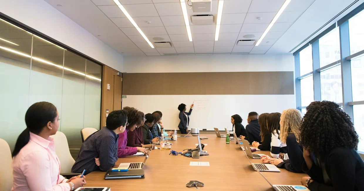 Colleagues seated around a long boardroom table listening to a woman speaking