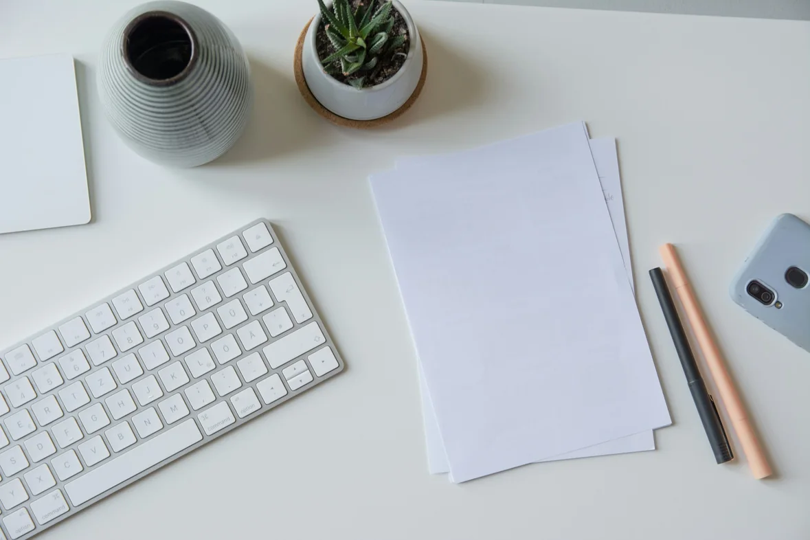 A tidy white desk with a keyboard, blank sheets of paper, a pen and a small plant