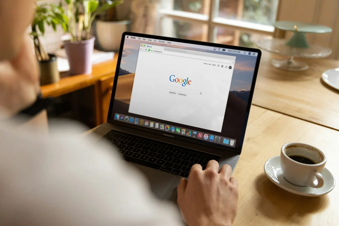 A laptop open on a wooden table showing the Google search page, a cup of coffee and plants beside it