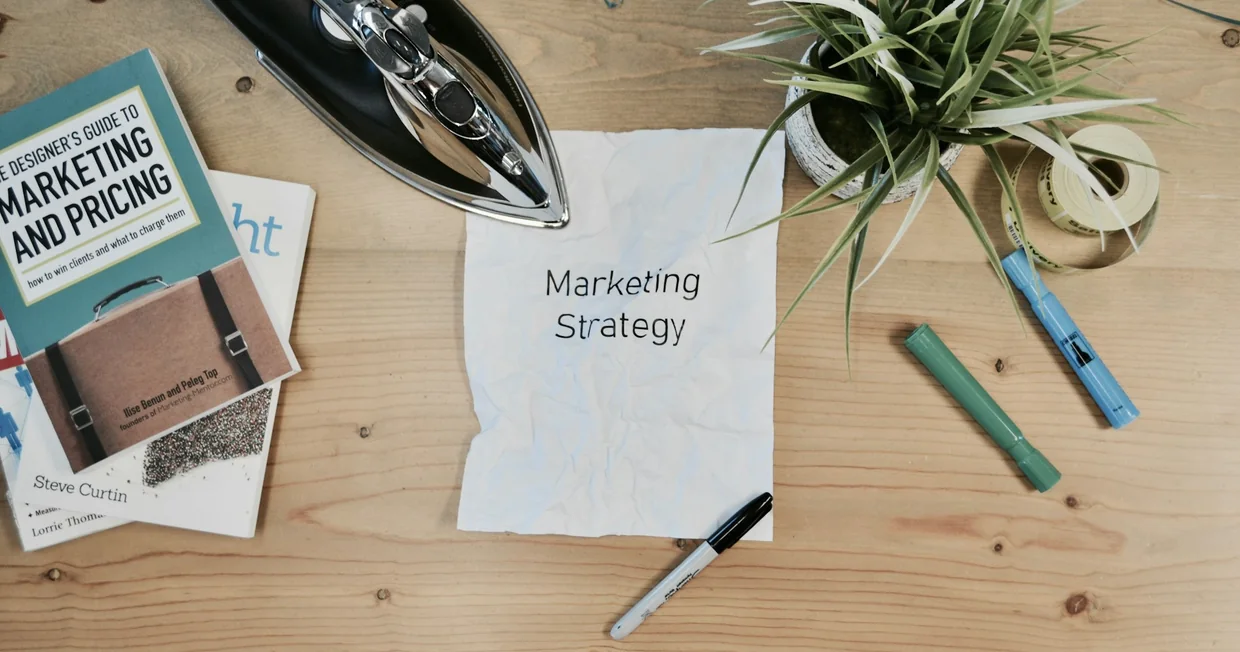 A light wooden desk from above: a handwritten note reading Marketing Strategy, a book on marketing and pricing, business cards, a pen, marker pens and a small plant