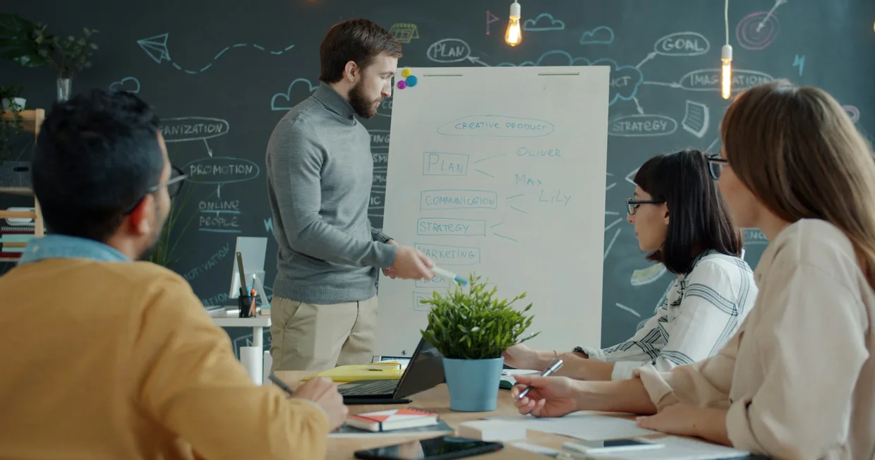 A team standing and sitting around a whiteboard in a bright office, one person writing on it while the others follow along with notebooks