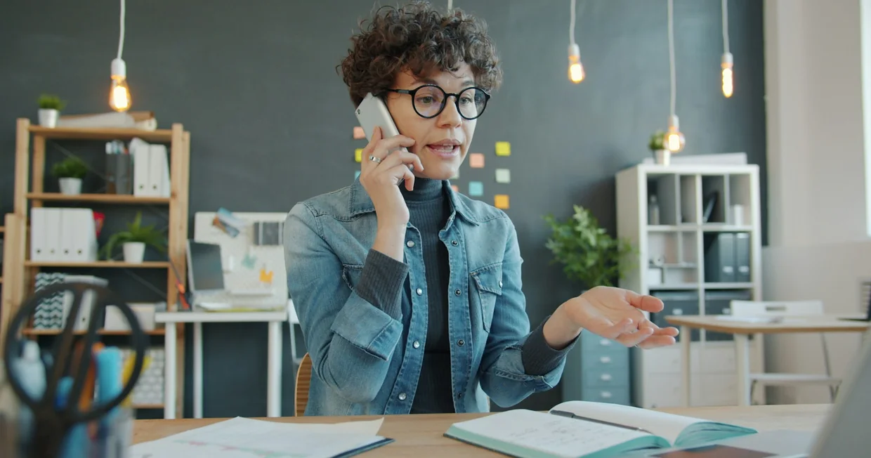 A person at an office desk speaking on a mobile phone, gesturing, with notebooks in front of them