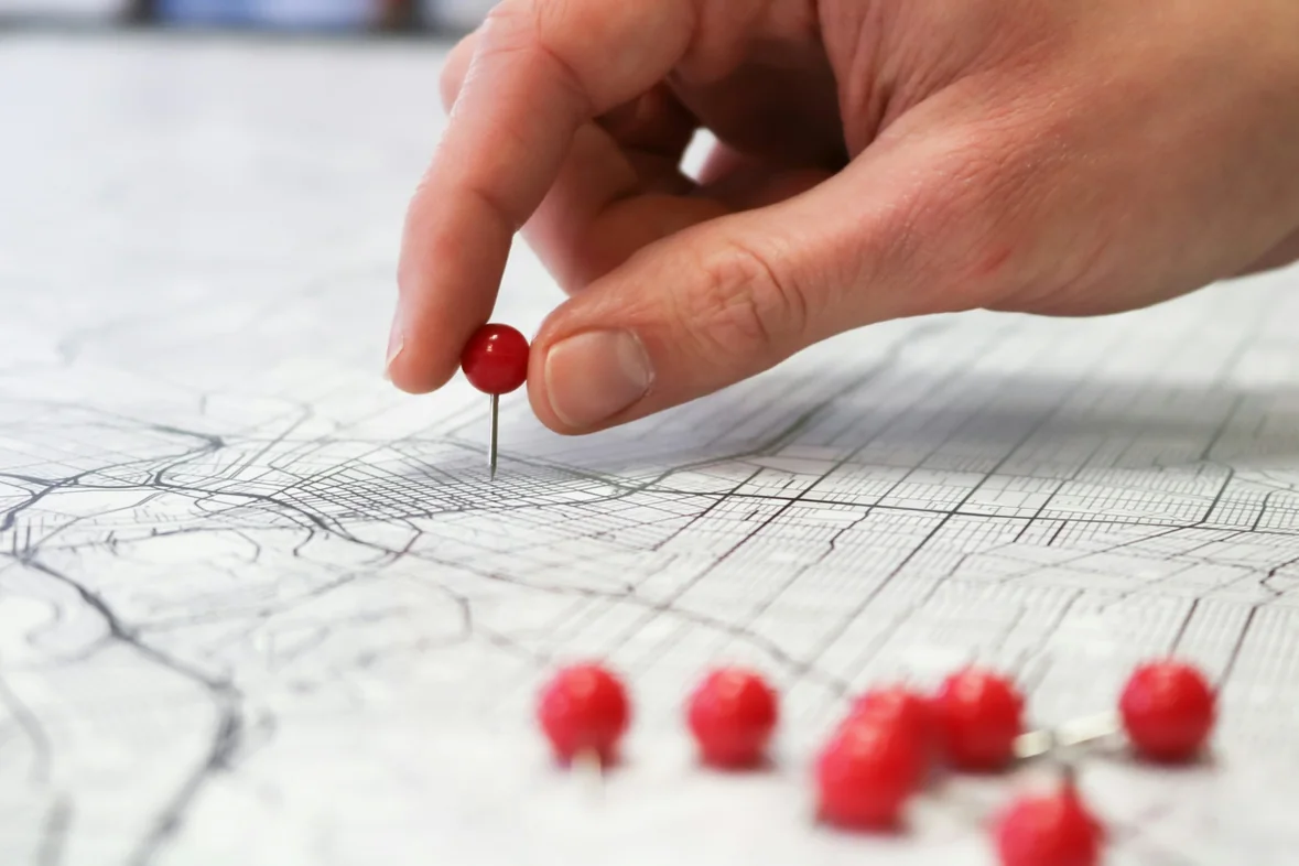 A hand pressing a red map pin into a printed street map, with several loose pins resting beside it
