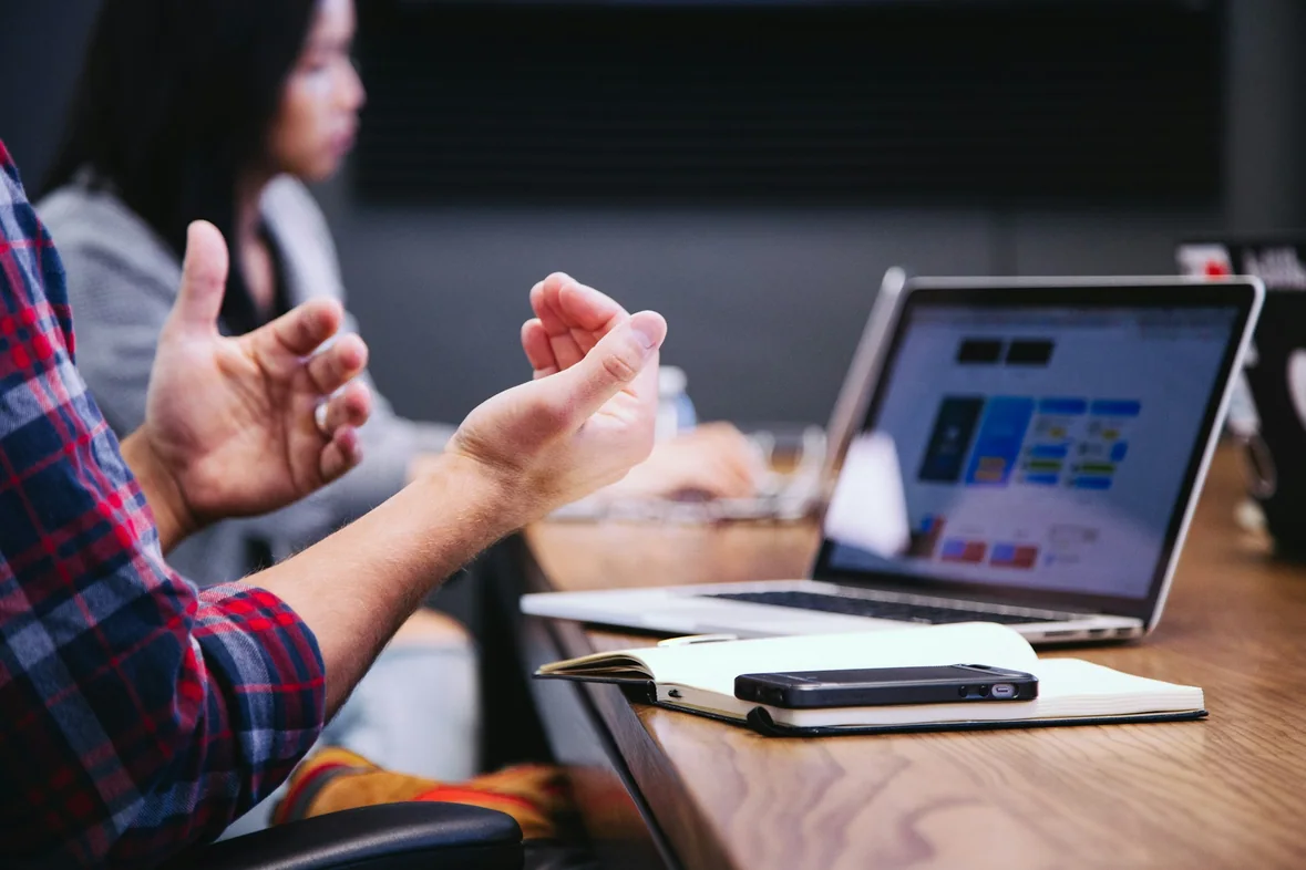 Someone gesturing while explaining something at a meeting table beside an open laptop