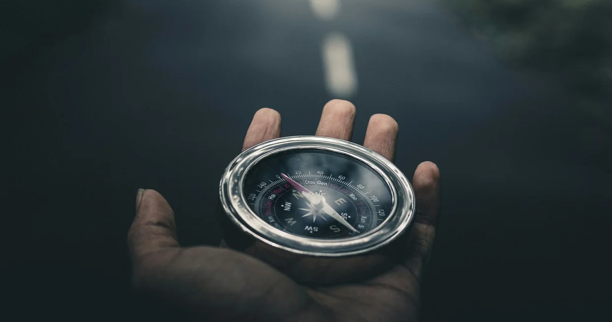 A hand holding a small silver compass over open ground