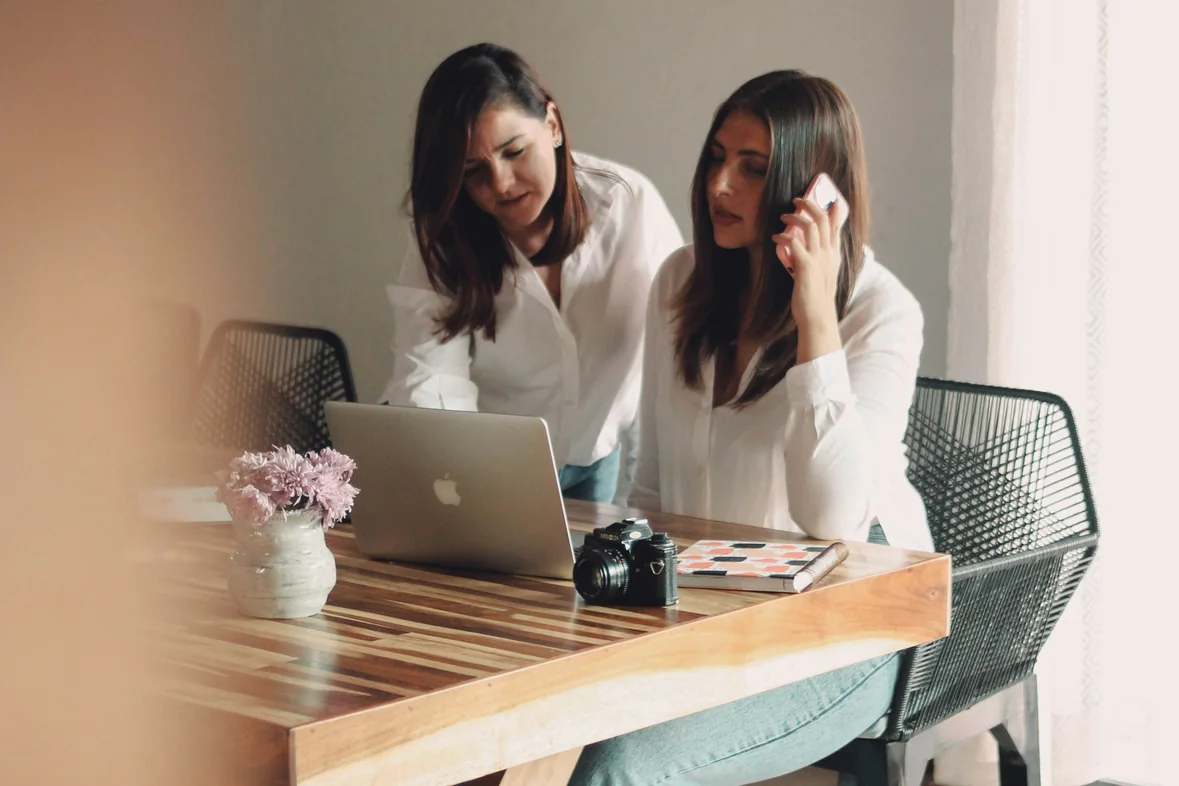 Two colleagues at a wooden table with an open laptop, one of them speaking on a mobile phone