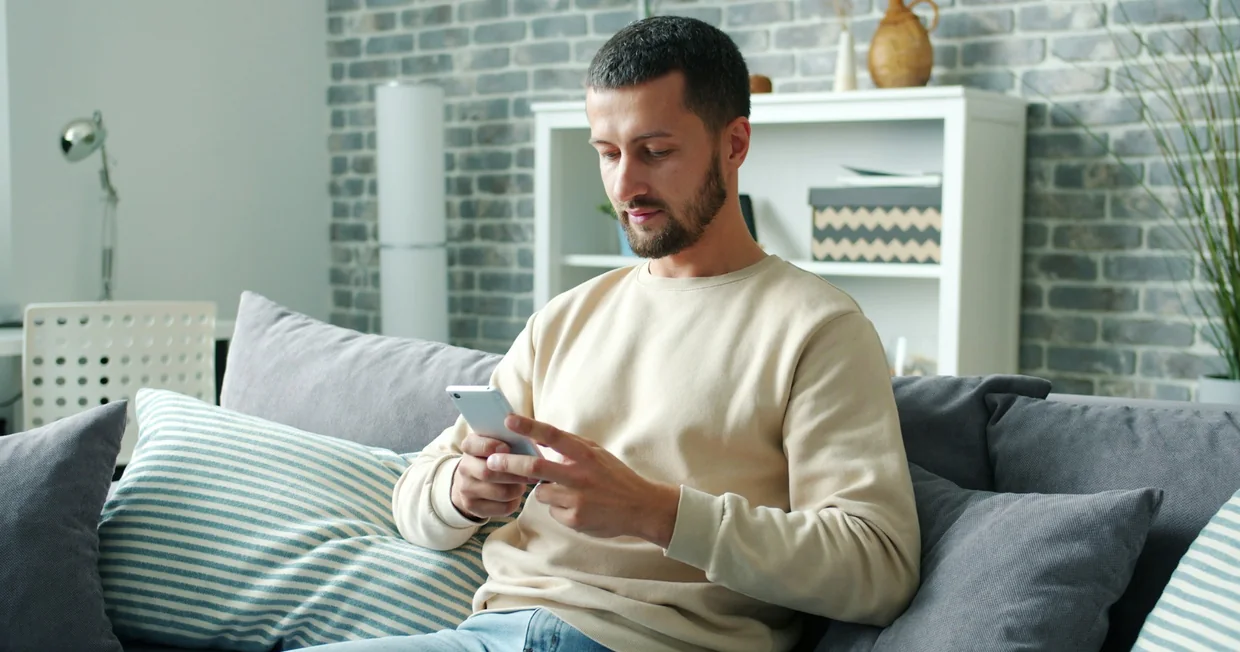 A man sitting on a sofa in a living room, looking down at the phone in his hands