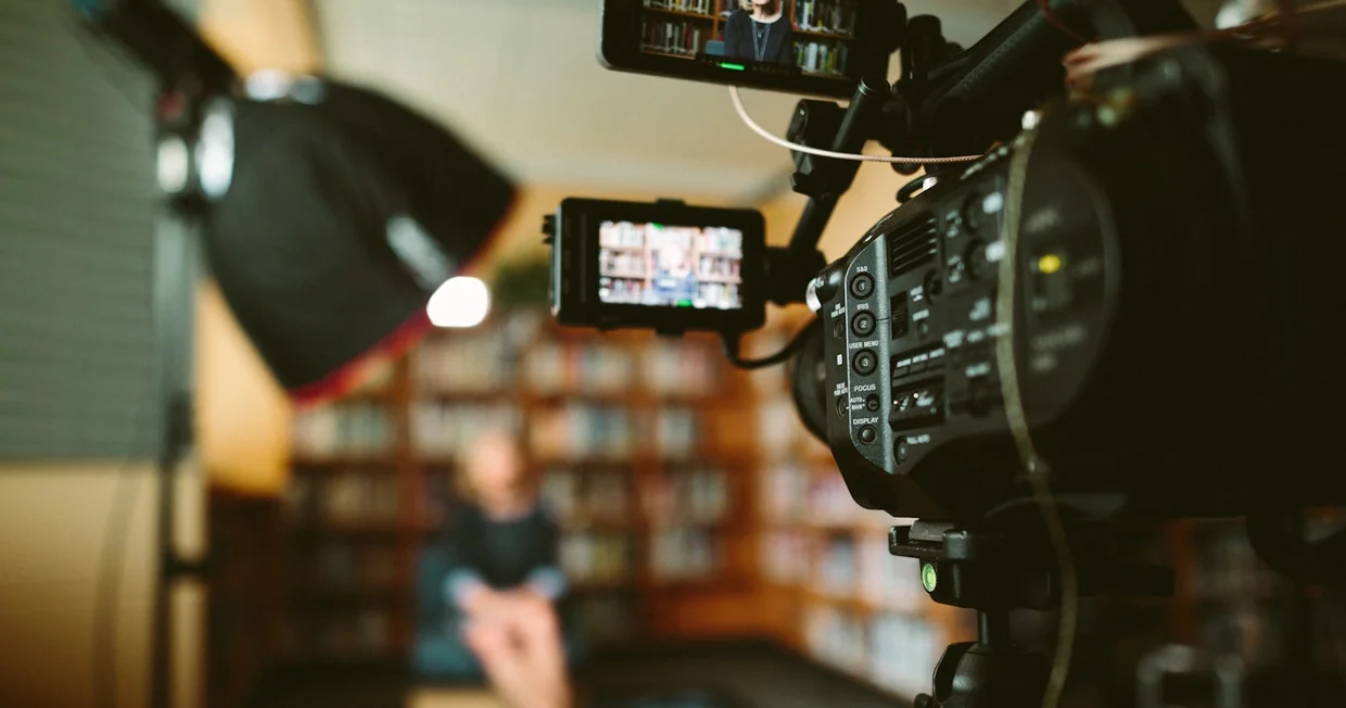 A person seated in front of a bookshelf being filmed