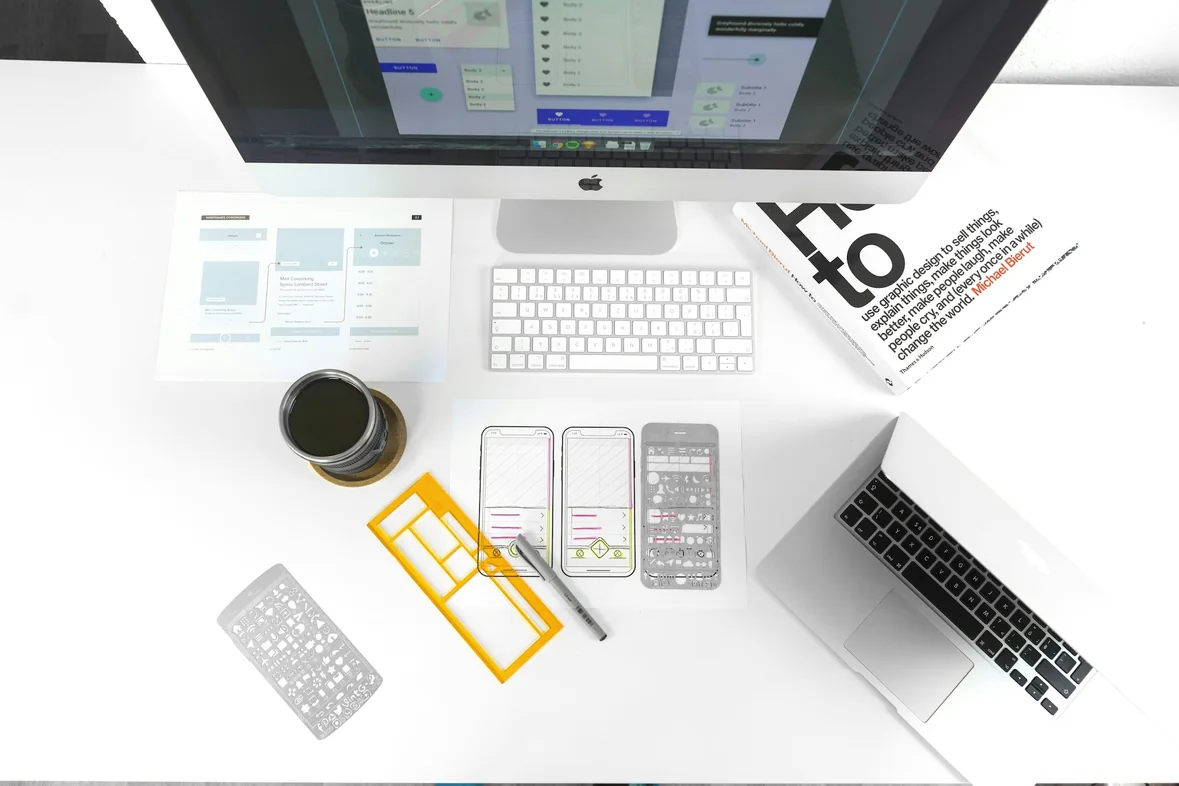 A tidy white desk with a monitor, keyboard, laptop, coffee cup and printed page-layout cards arranged beside them