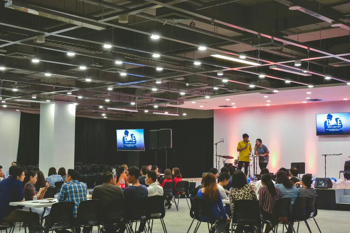 An audience seated at tables in a large hall watching a speaker present from a lit stage