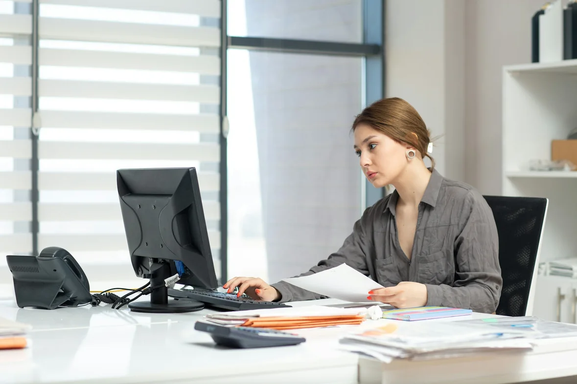 A person at an office desk working through printed documents beside a computer monitor